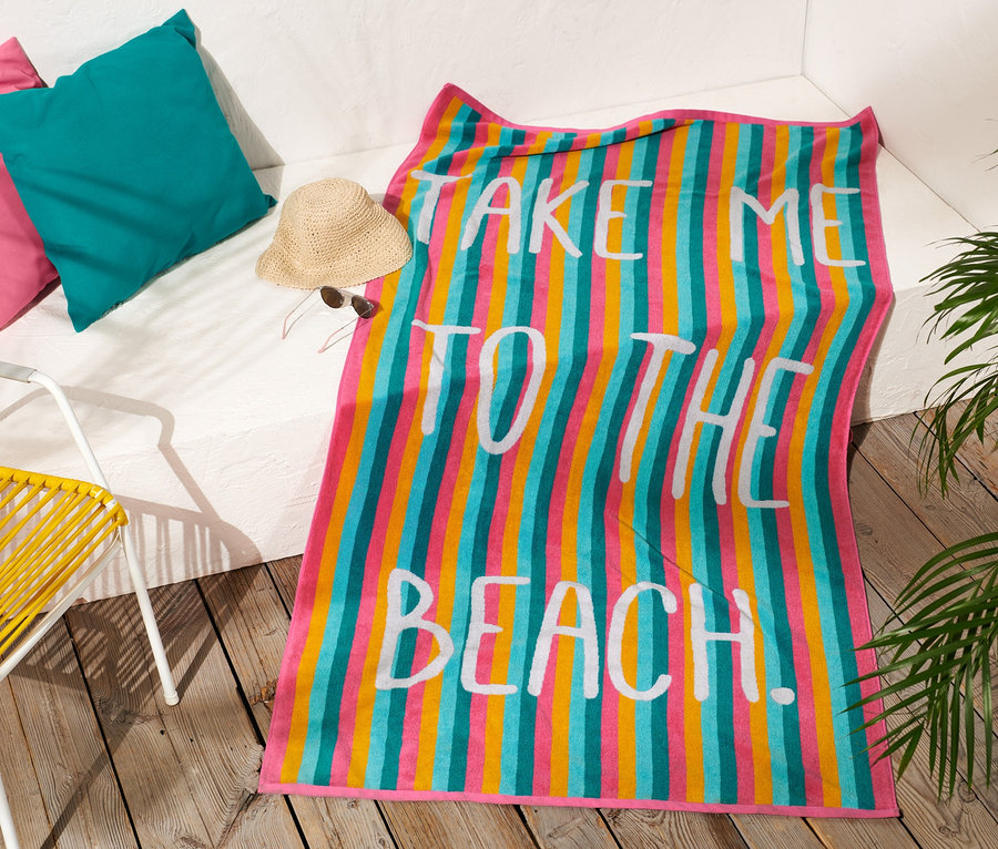 Ein bunt gestreiftes XL-Frottier-Badetuch mit dem Aufdruck "Take me to the beach" liegt auf einem Holzboden neben einem gelben Stuhl.