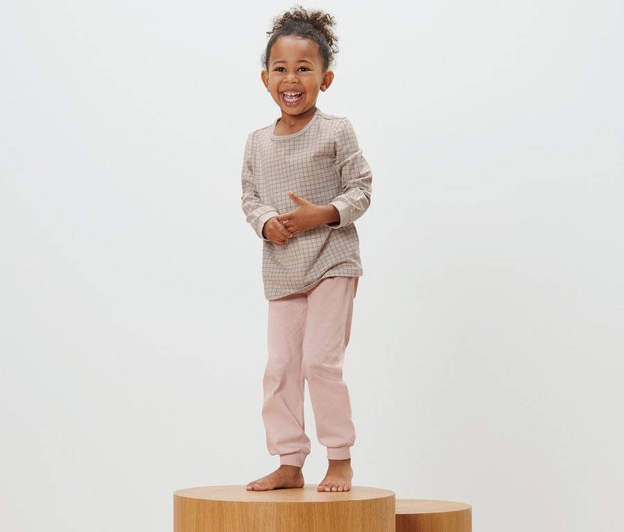 Lächelndes Mädchen steht auf einem Holzsockel und trägt einen rosa, langen Kinder-Pyjama.