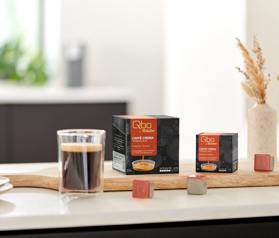 Ein Glas Kaffee steht auf einem Holzbrett neben zwei schwarzen Packungen Qbo Caffè TERRENO ALTO Kapseln, eine mit 27 und eine mit 8 Kapseln. Zwei einzelne Qbo Kapseln, eine rotbraune und eine graue, liegen vor den Packungen. Rechts steht eine Vase mit Trockengras, links eine Schale mit Keksen.