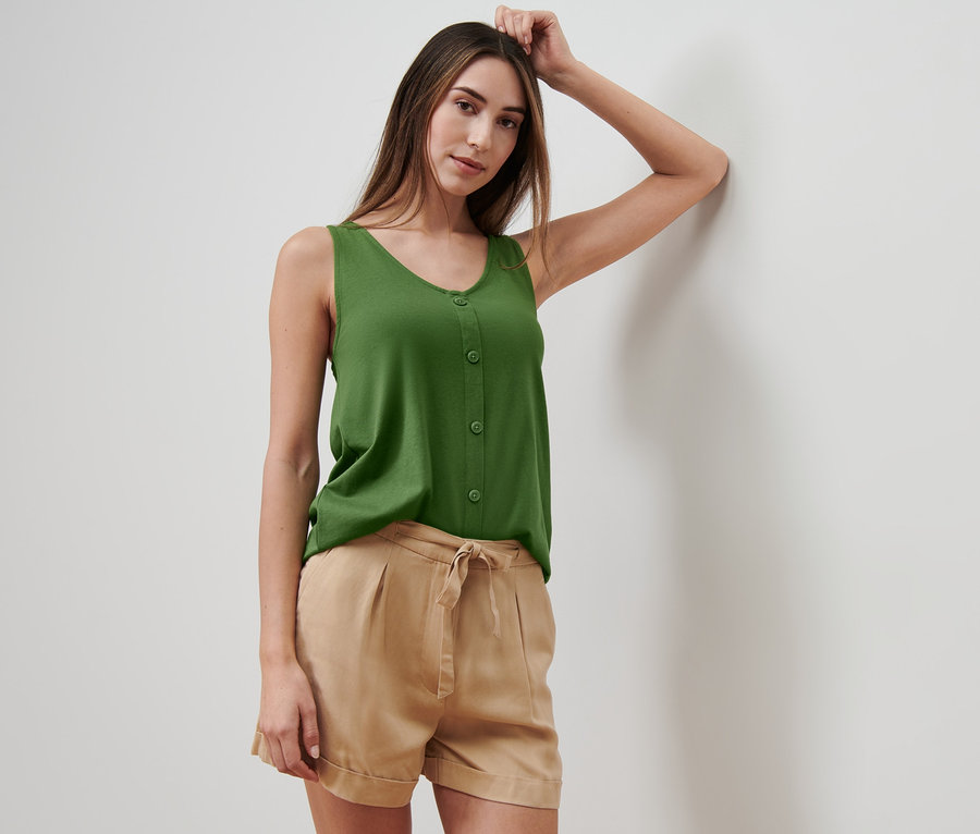Frau an eine Wand gelehnt trägt ein grünes Top mit Knopfleiste und eine beige Shorts.