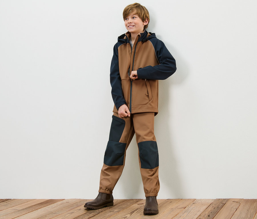Junge posiert in brauner Softshelljacke und Kinder-Softshellhose vor einer weißen Wand.