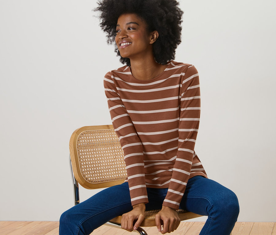 Frau sitzt auf einem Stuhl und trägt ein gestreiftes Langarmshirt in Cognac.