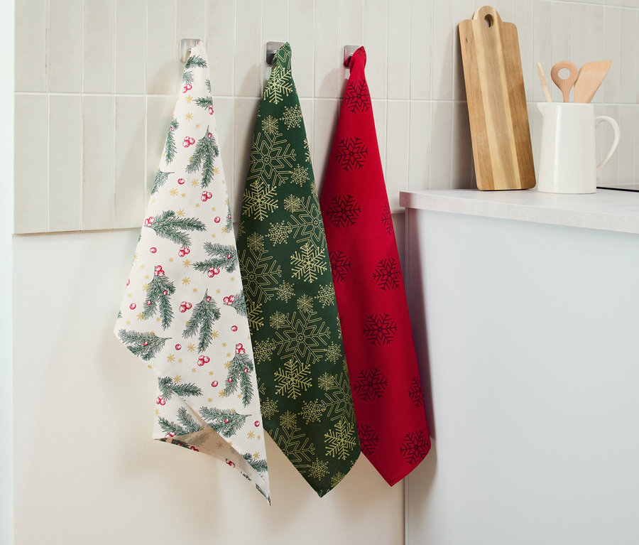 Drei Geschirrtücher hängen an der Wand: ein weißes mit Weihnachtsmotiv, ein grünes mit goldenen Schneeflocken und ein rotes mit dunklen Schneeflocken.