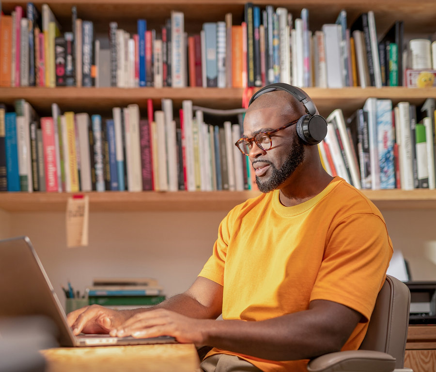 Mann mit Brille und Philips Bluetooth®-Kopfhörern »H3209« tippt auf einem Laptop. Im Hintergrund ein Bücherregal.
