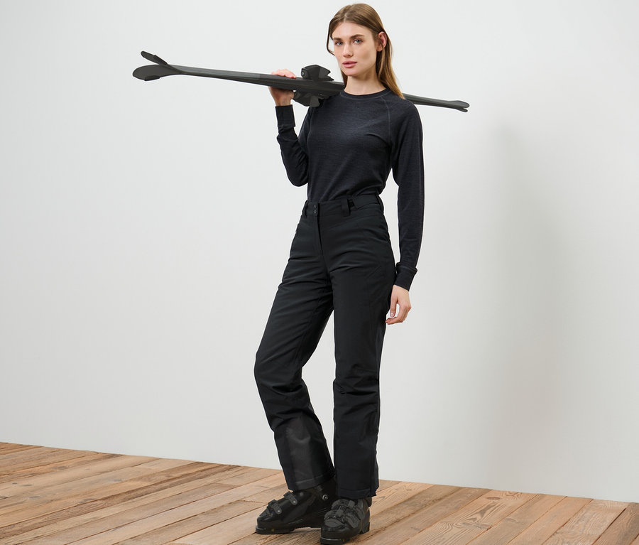 Frau posiert in Skihose und hält Ski über die Schulter.