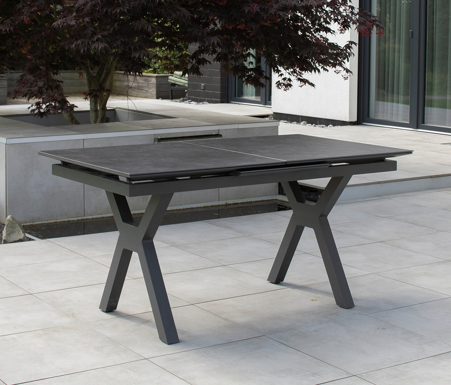 Ein steingrauer deVries Alu Ausziehtisch, 160/210, steht auf einer Terrasse neben einem Baum.