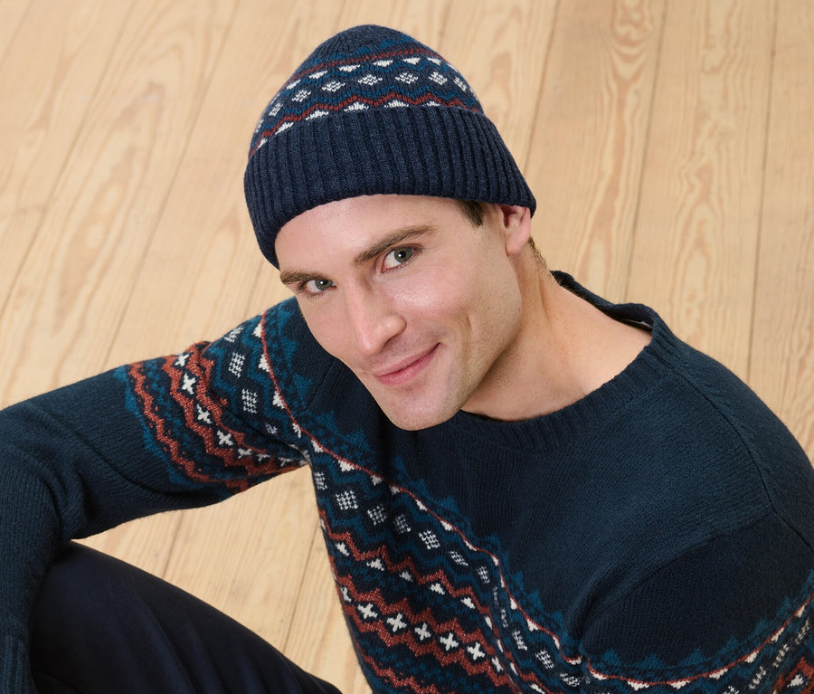 Ein Mann sitzt auf einem Holzboden und trägt eine Strickmütze mit RWS-zertifizierter Wolle und einen Pullover mit Muster.