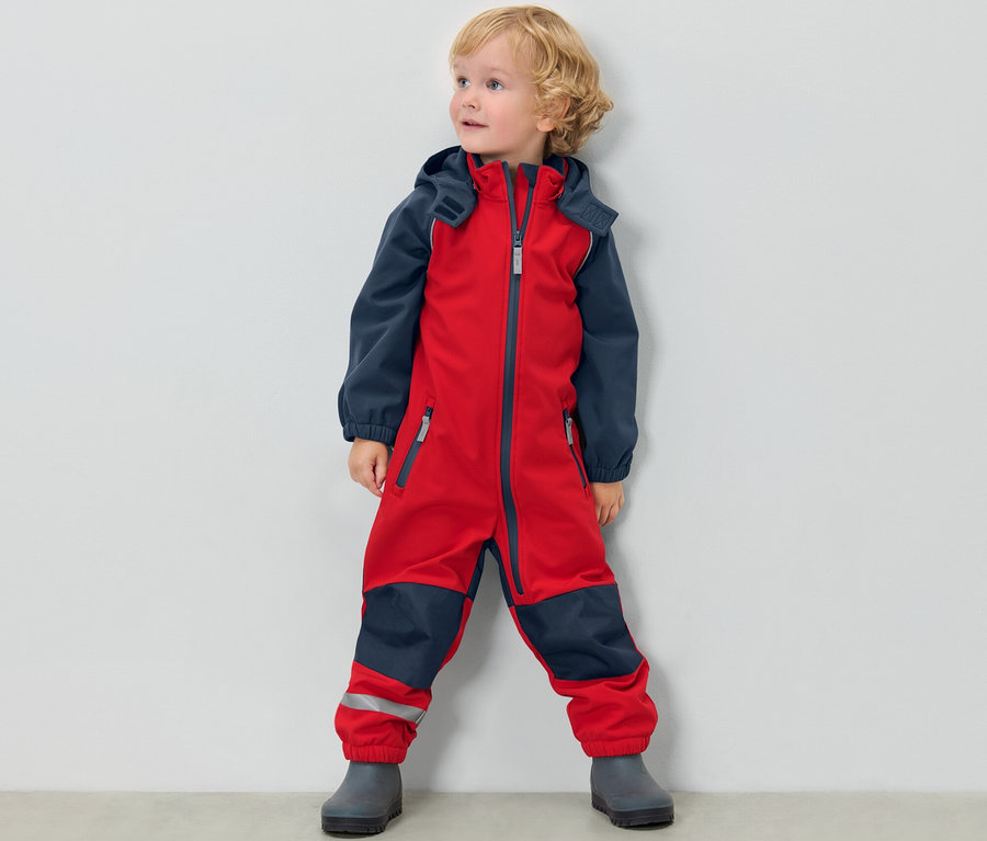 Kleiner Junge steht in rot-blauem Kinder-Softshell-Einteiler vor einer weißen Wand.