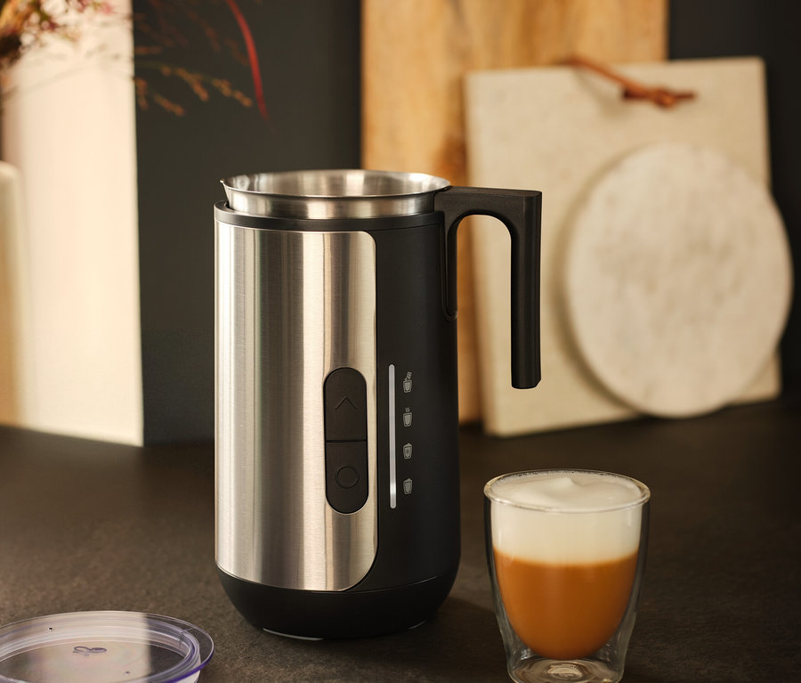 Ein Milchaufschäumer aus Edelstahl steht neben einem Glas Kaffee mit Milchschaum.
