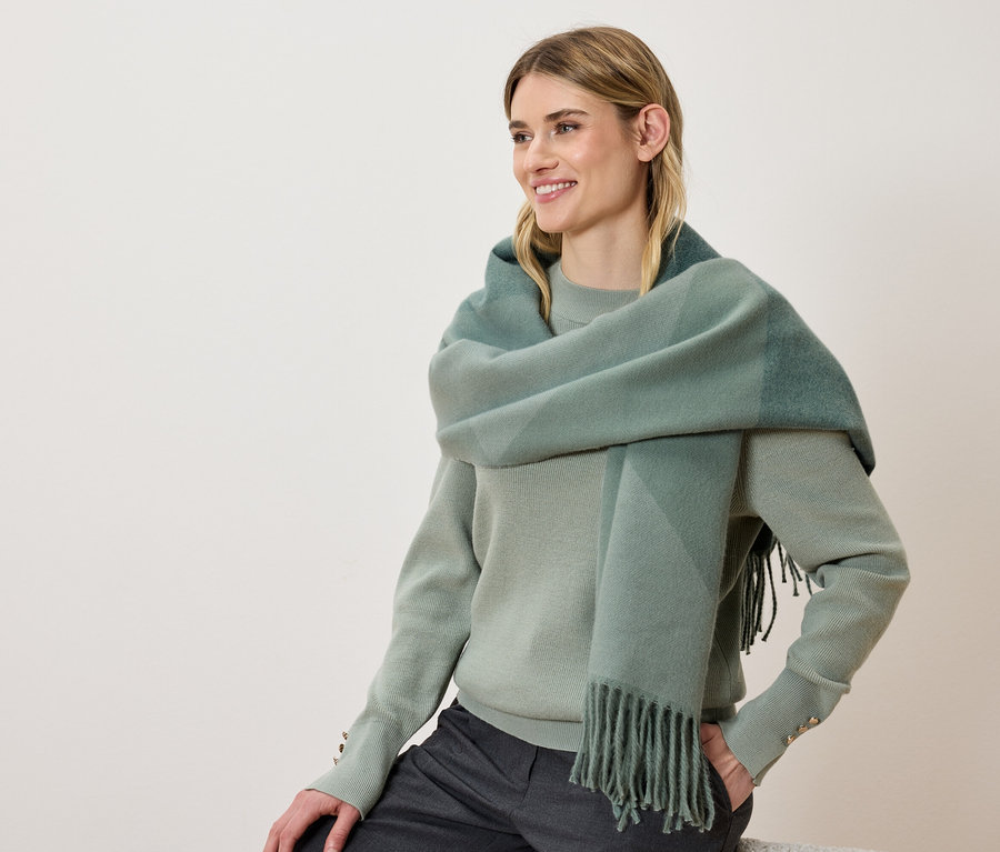 Eine lächelnde Frau trägt einen grünen Pullover und einen karierten Schal mit Fransen.
