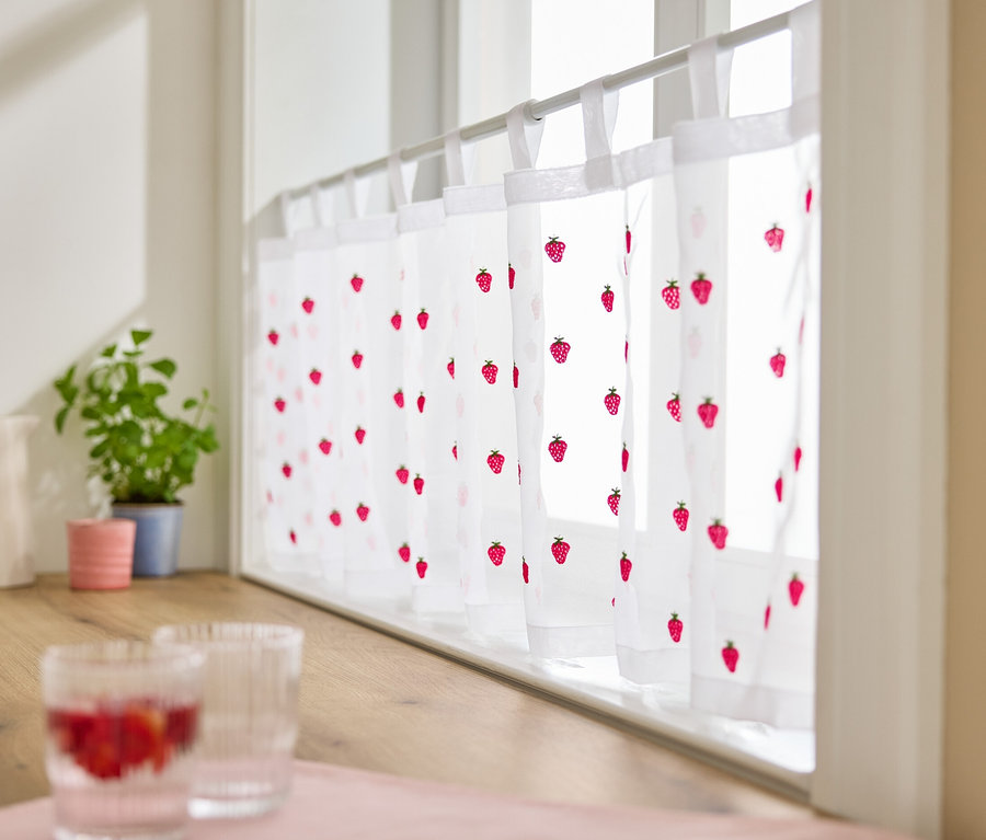 Am Fenster hängt eine bestickte Bistrogardine mit Erdbeermuster.