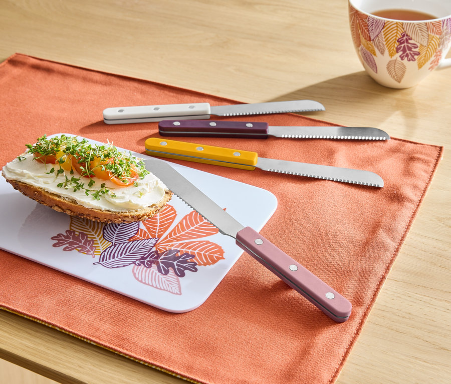 Auf einem orangefarbenen Tischset liegt ein Brot mit Aufstrich auf einem Brettchen mit Blättermuster, daneben liegen vier Messer mit bunten Griffen und im Hintergrund steht eine Tasse Tee.