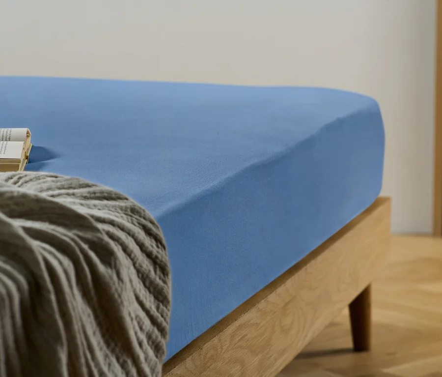 Detailaufnahme eines blauen Spannbetttuchs auf einem Bett mit einem Buch und einer grauen Decke.