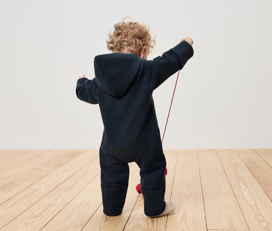 Kleinkind im blauen Overall spielt mit einem roten Spielzeug an einer Schnur.