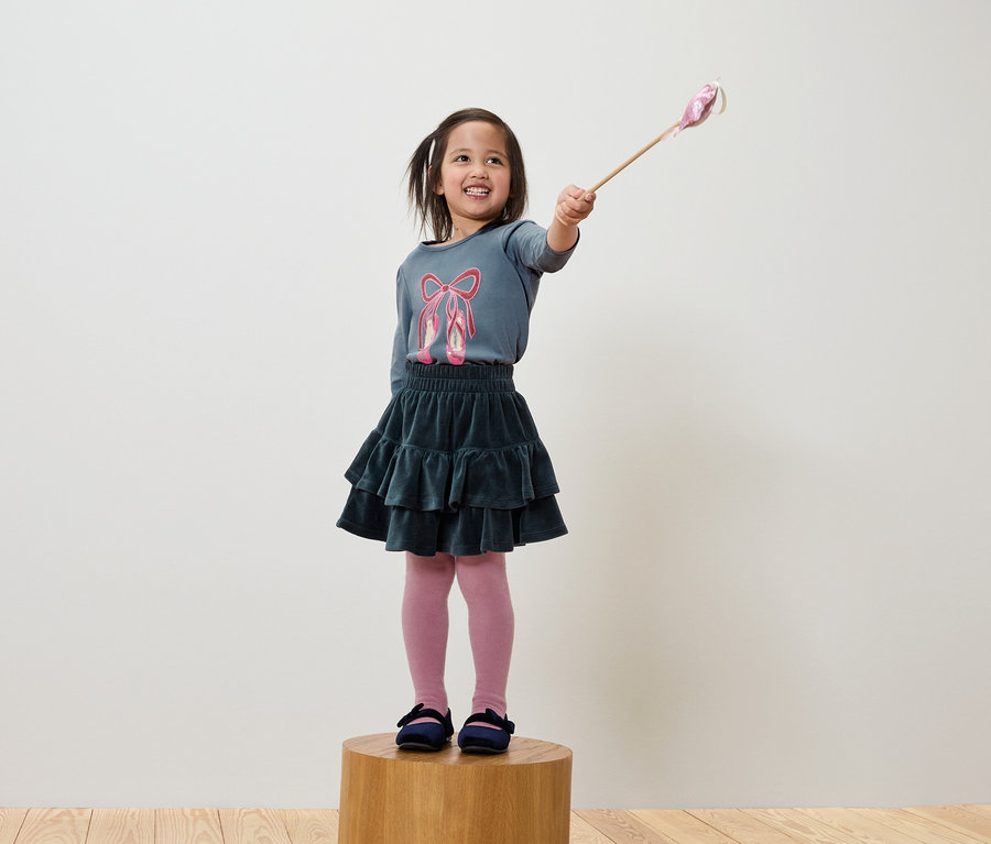 Ein lächelndes Mädchen hält einen Zauberstab und trägt ein blaues T-Shirt mit Ballettschuhen, einen grünen Rock und eine rosa Strumpfhose.