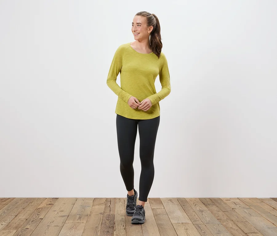 Frau in einem gelbgrünen Langarmshirt, schwarzen Leggings und grauen Schuhen.
