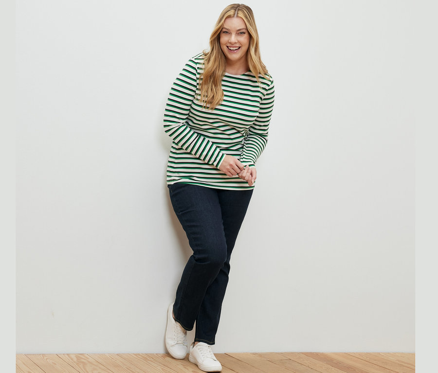 Eine lächelnde Frau lehnt an einer Wand und trägt ein mehrfarbig gestreiftes Langarmshirt und eine dunkelblaue Straight Jeans – Fit »Kira«.