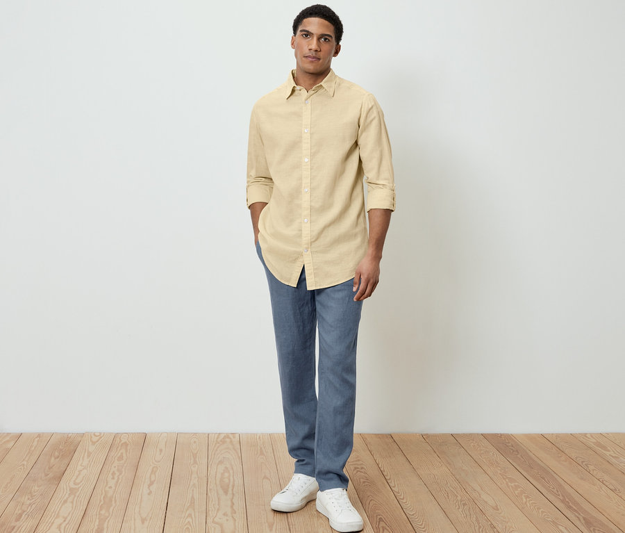 Mann posiert in gelbem Leinenhemd, blauen Hosen und weißen Schuhen.