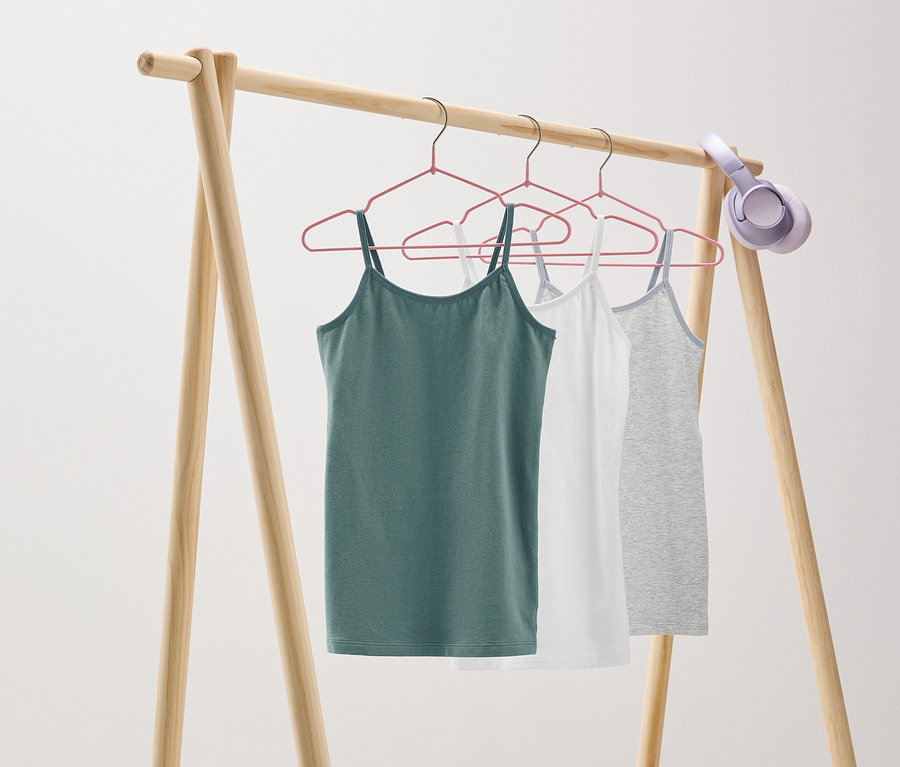 Auf einem hölzernen Kleiderständer hängen drei Tanktops in Grün, Weiß und Grau an rosa Kleiderbügeln. Am Ständer hängt ein Kopfhörer.
