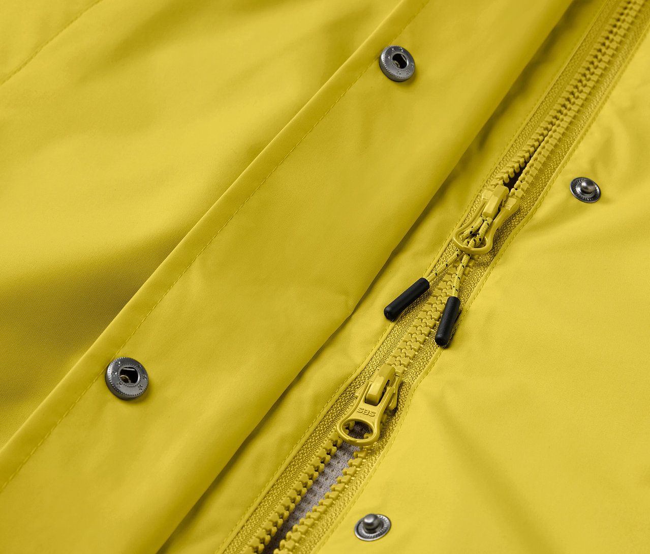 Detailaufnahme einer gelben Jacke mit silbernen Druckknöpfen und gelbem Reißverschluss.