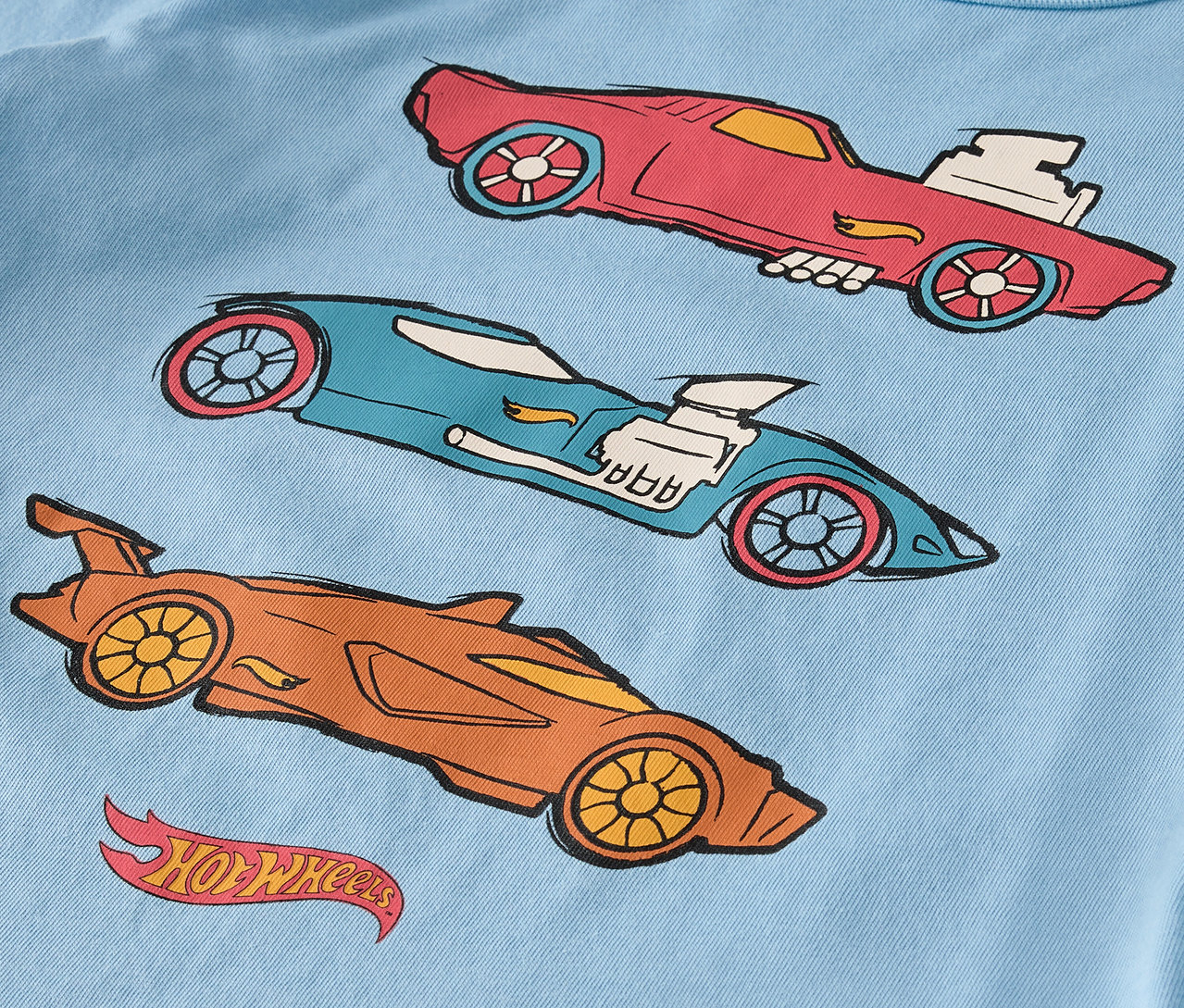 Auf einem hellblauen T-Shirt sind drei gezeichnete Hot Wheels Autos in Rot, Blau und Orange.