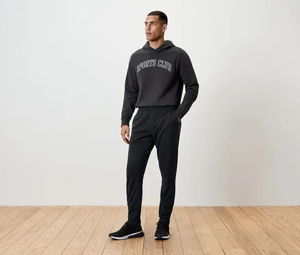 Mann steht auf einem Holzboden und trägt eine schwarze Trainingshose, einen Kapuzenpullover und Turnschuhe.