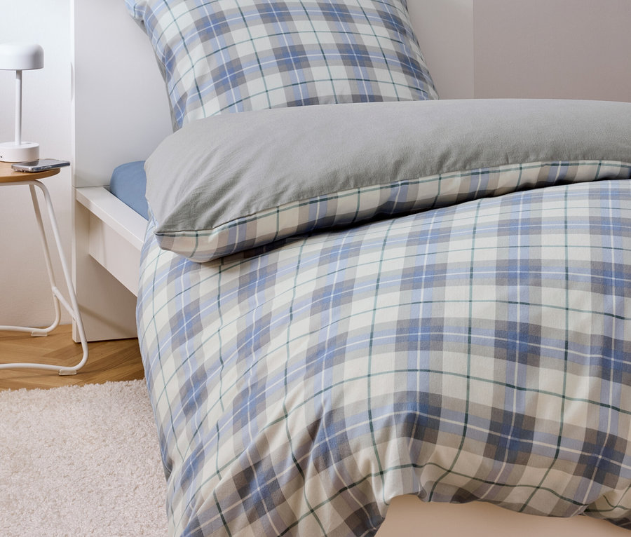 Bett mit blau-weiß karierter Bettwäsche und grauem Kissen.