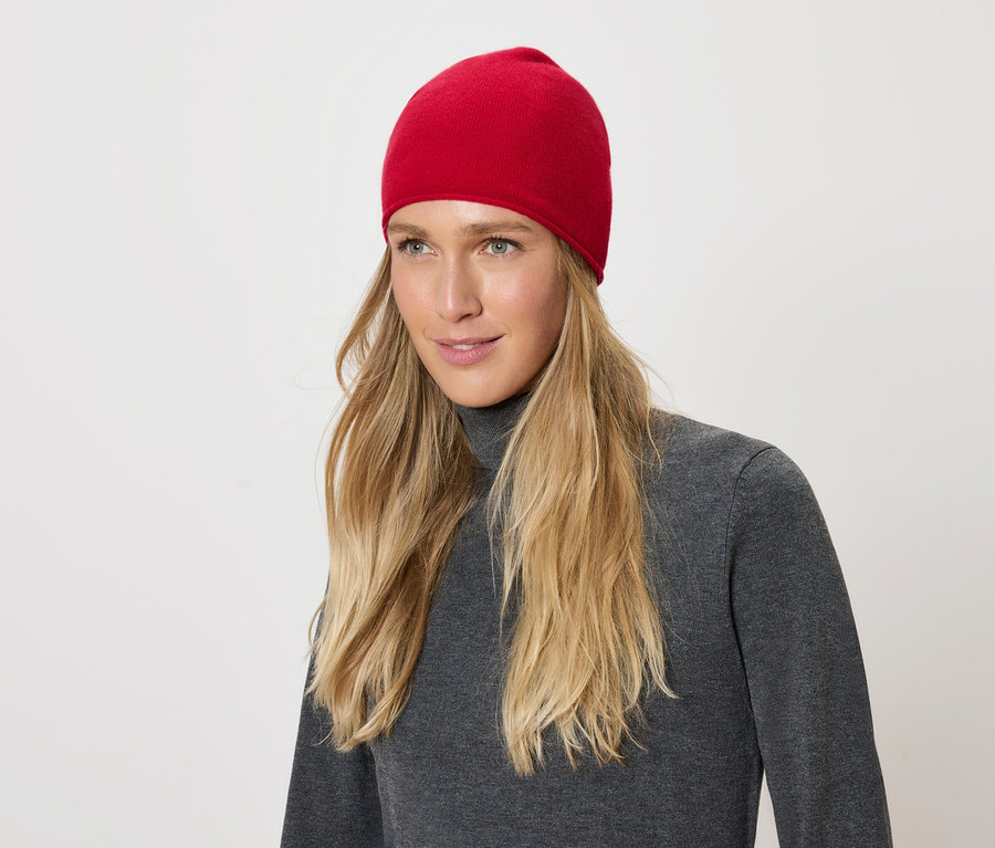 Porträt einer Frau mit blonden Haaren und einer roten Mütze, die einen grauen Rollkragenpullover auf weißem Hintergrund trägt.