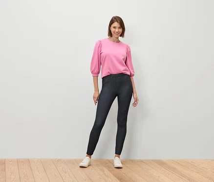 Eine Frau steht auf einem Holzboden und trägt eine rosa Bluse und dunkelblaue Jeans.
