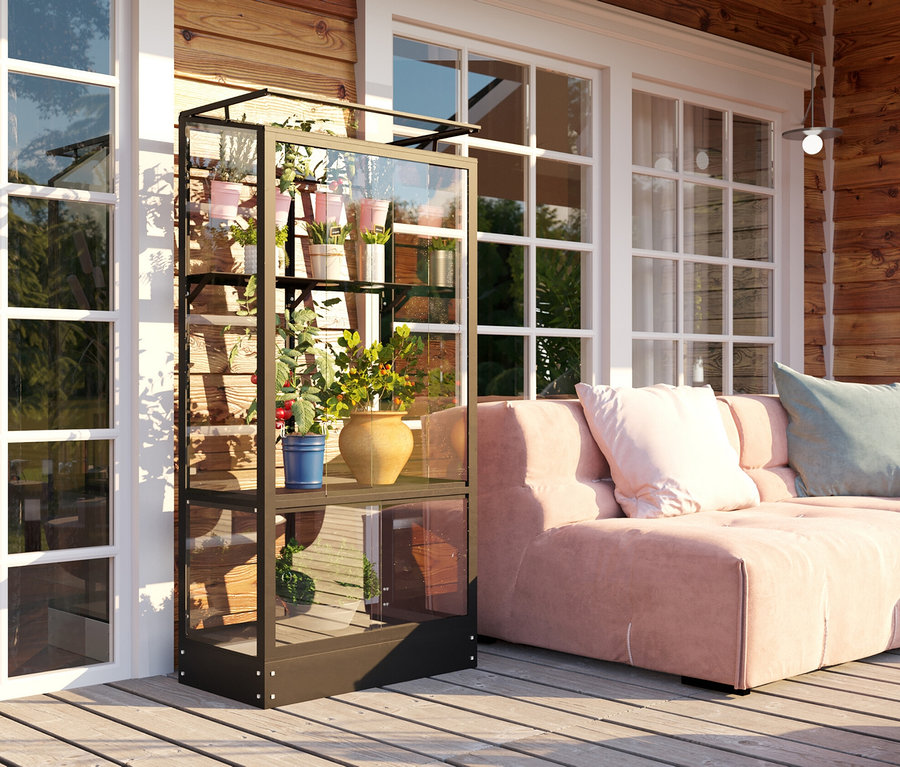 Ein schwarzes Vitavia Balkongewächshaus mit Standfundament steht auf einer Holzterrasse neben einem rosa Sofa.