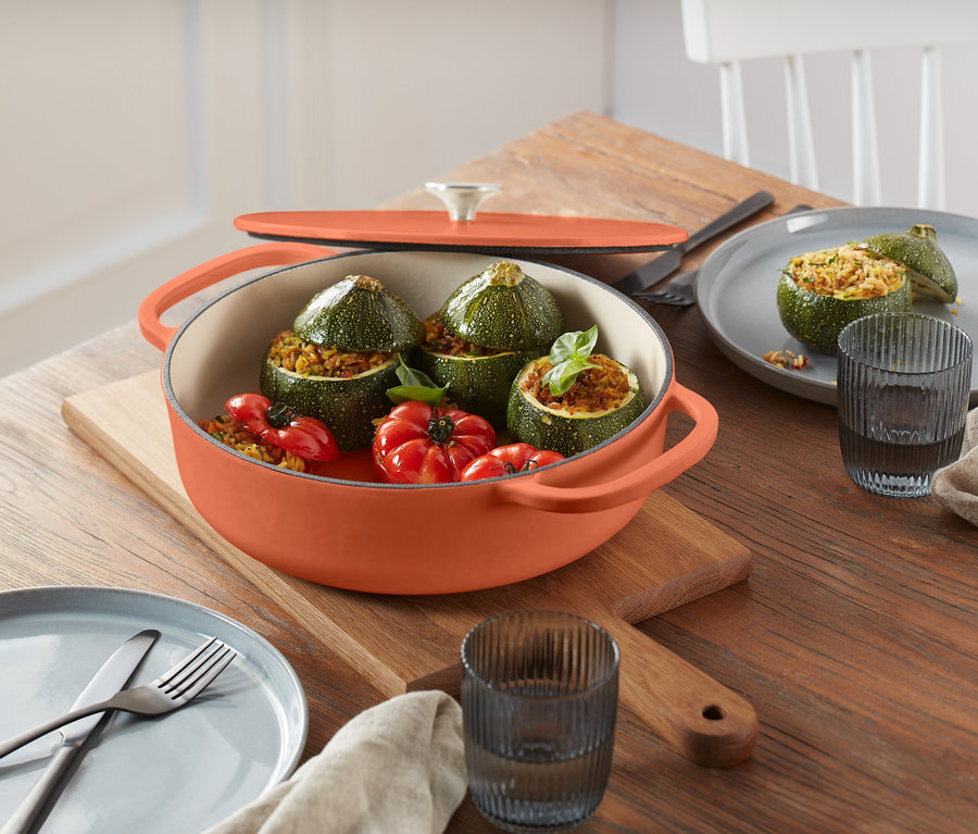 Gefüllte Zucchini und Tomaten in einem orangefarbenen Topf auf einem Holzbrett.