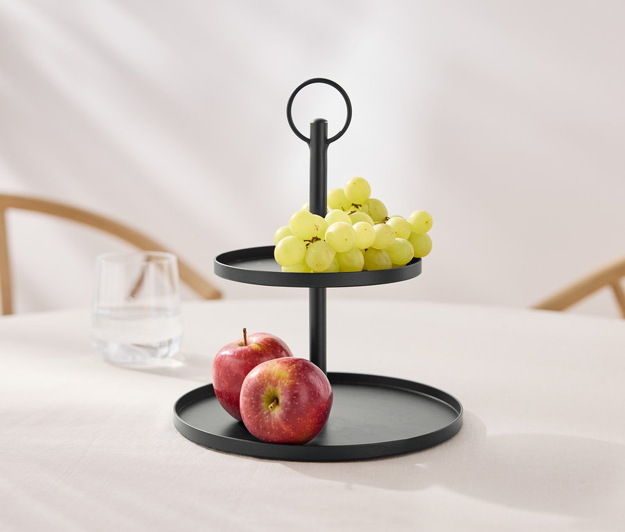 Ein zweistöckiger, schwarzer Etagere mit Trauben und Äpfeln auf einem Tisch.