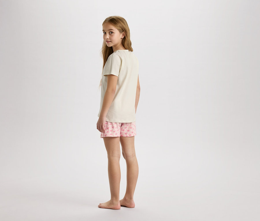 Ein junges Mädchen steht barfuß in einem beigen T-Shirt und rosa Shorts mit Blumendruck vor einem neutralen Hintergrund.