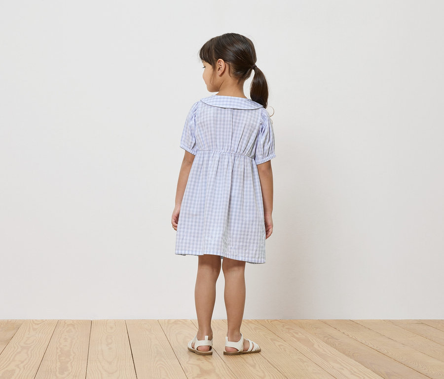 Ein junges Mädchen steht mit dem Rücken zur Kamera und trägt ein blau-weiß kariertes Kleid und Sandalen.