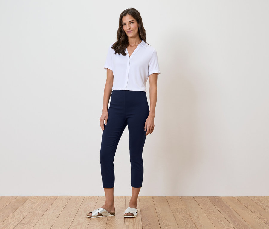 Frau steht vor einer Wand und trägt ein weißes Hemd, blaue Hosen und goldene Sandalen.