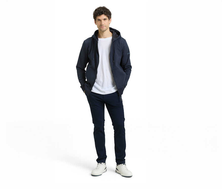 Mann in dunkelblauer Kapuzenjacke über weißem T-Shirt, dunkelblauer Hose und weißen Sneakers, Hände in den Hosentaschen.