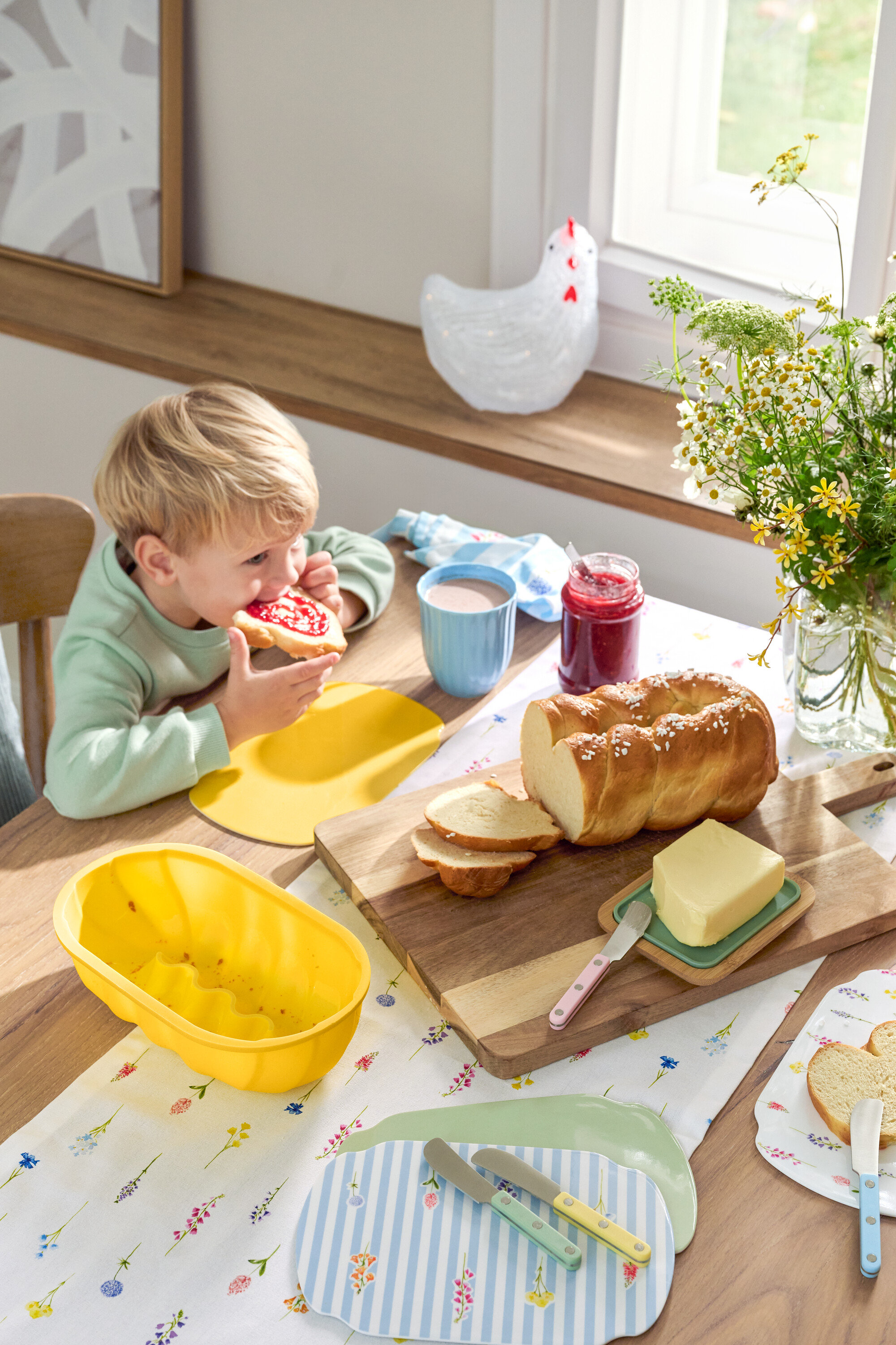 Blonder Junge isst ein Marmeladenbrot am Tisch mit Striezel, Butter, Marmelade, Geschirr und Blumen.