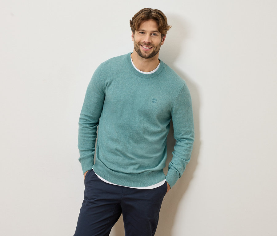 Mann mit hellbraunem Haar und Bart, der einen hellblauen Pullover und eine marineblaue Hose trägt und lächelt.