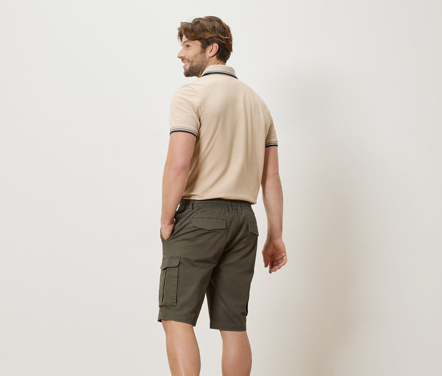 Mann von hinten in beigem Poloshirt und grünen Shorts mit Händen in den Taschen.