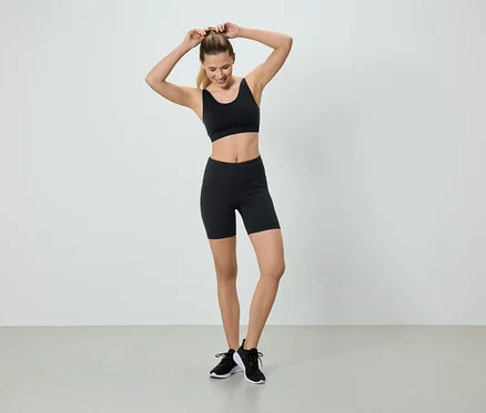 Eine Frau mit blonden Haaren, die einen schwarzen Sport-BH und Shorts trägt, steht vor einem weißen Hintergrund und macht sich einen Pferdeschwanz.