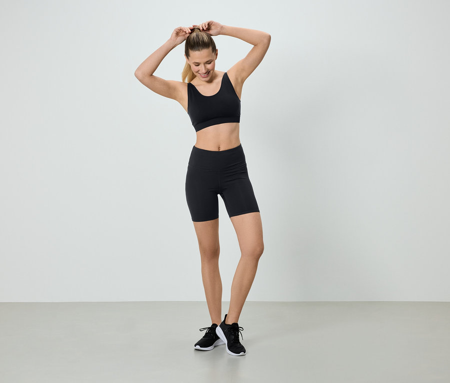 Eine Frau mit blonden Haaren, die einen schwarzen Sport-BH und Shorts trägt, steht vor einem weißen Hintergrund und macht sich einen Pferdeschwanz.