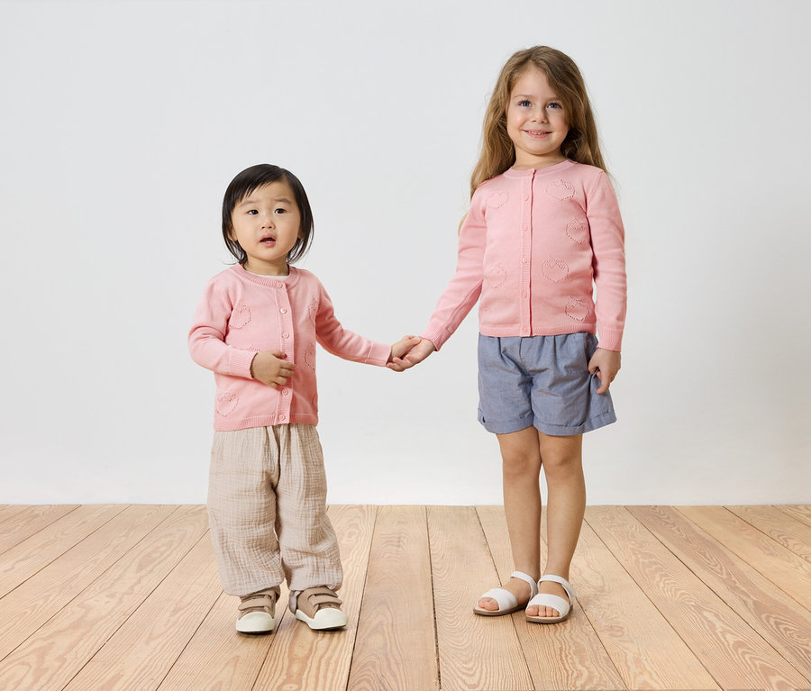 Zwei Mädchen, eines mit dunklen Haaren und eines mit blonden Haaren, halten Händchen. Beide tragen rosa Strickjacken, wobei das kleinere Mädchen dazu noch beige Hosen und Sneaker trägt und das größere blaue Shorts und Sandalen.
