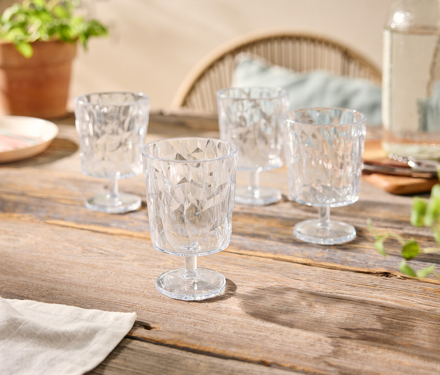 Vier klare, facettierte Gläser auf einem Holztisch, zusammen mit einer Wasserflasche, einem Teller und Topfpflanzen.