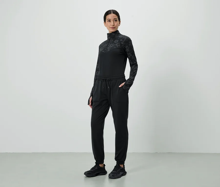 Frau in schwarzem, gemustertem Oberteil und Jogginghose mit Händen in den Taschen, dazu schwarze Turnschuhe.