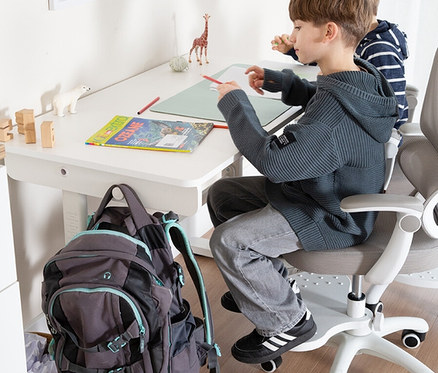 Zwei Kinder sitzen am weißen, höhenverstellbaren Roba Kinderschreibtisch Basic ohne Schublade und malen. Ein Rucksack hängt am Tisch.