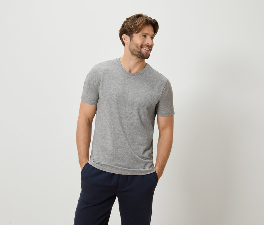 Mann mit grauem T-Shirt und blauen Hosen, die Hände in den Hosentaschen vor einem weißen Hintergrund.