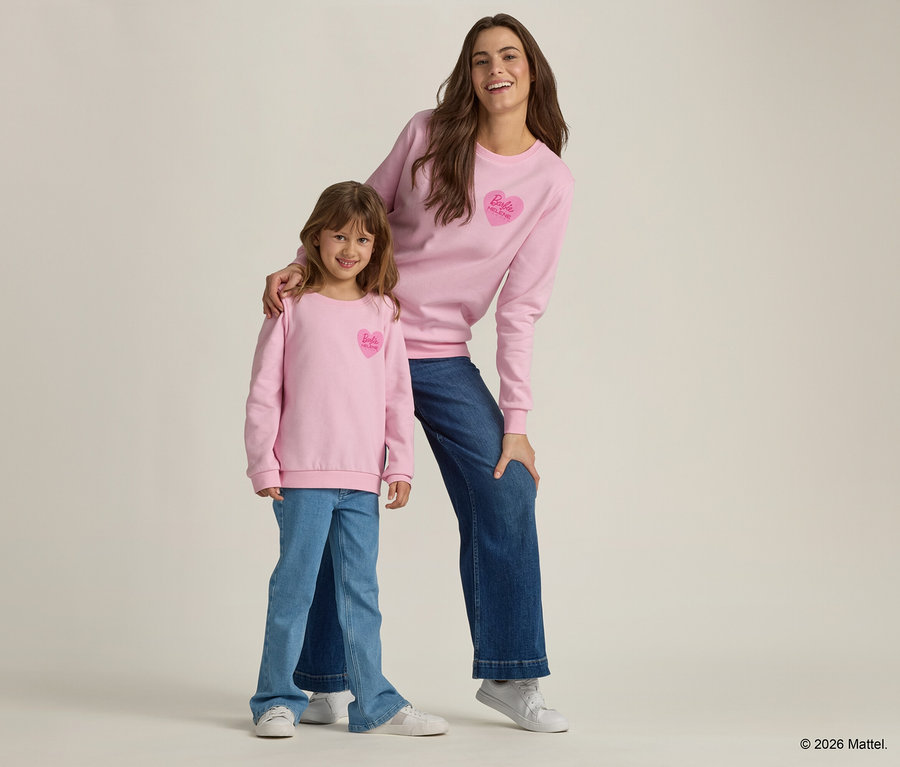 Eine Frau und ein Mädchen stehen zusammen und tragen rosa Pullover mit einem Barbie-Herz-Logo und Jeans.