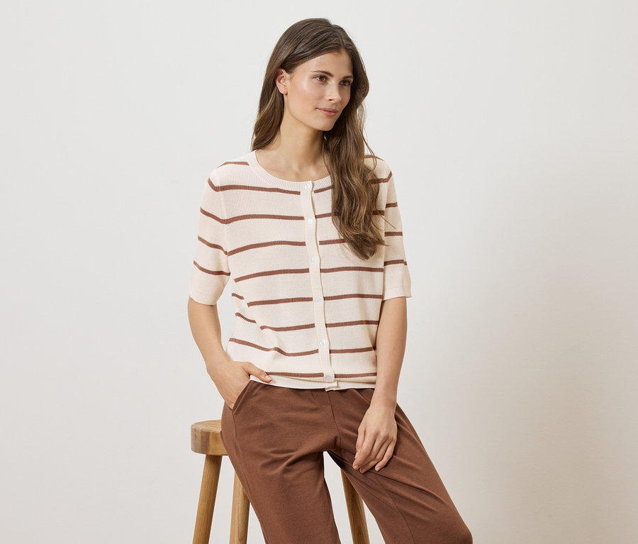 Frau sitzt auf einem Hocker, trägt eine gestreifte, kurzärmelige Strickjacke und braune Hose.