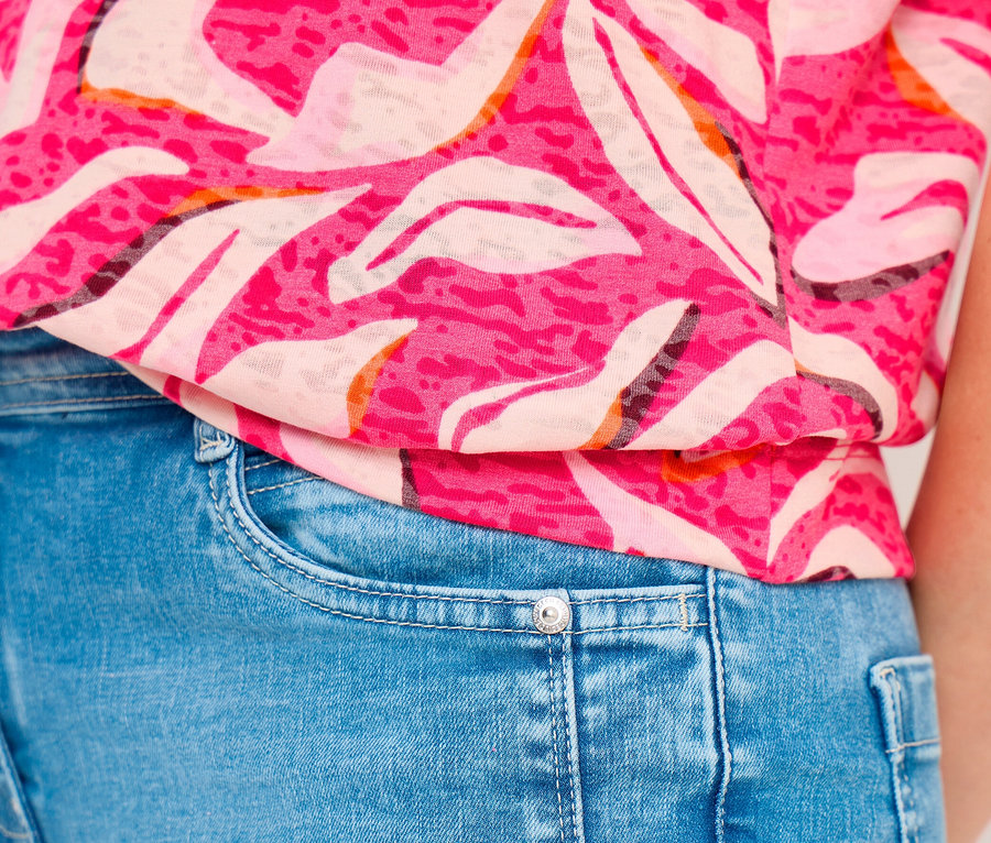 Nahaufnahme einer Person in einem pinkfarbenen CECIL Shirt mit weißen und orangen abstrakten Mustern und hochgekrempelten Ärmeln, das in hellblaue Jeans gesteckt ist.