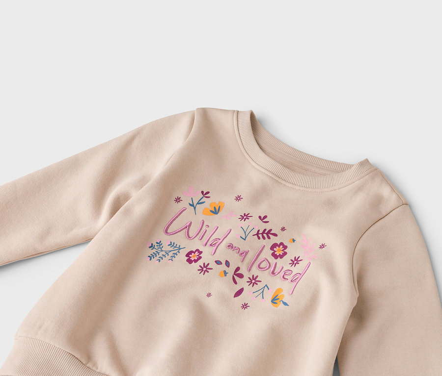 Nahaufnahme eines beigen Sweatshirts mit der Aufschrift "Wild and loved" und Blumen in Rosa, Lila und Gelb.